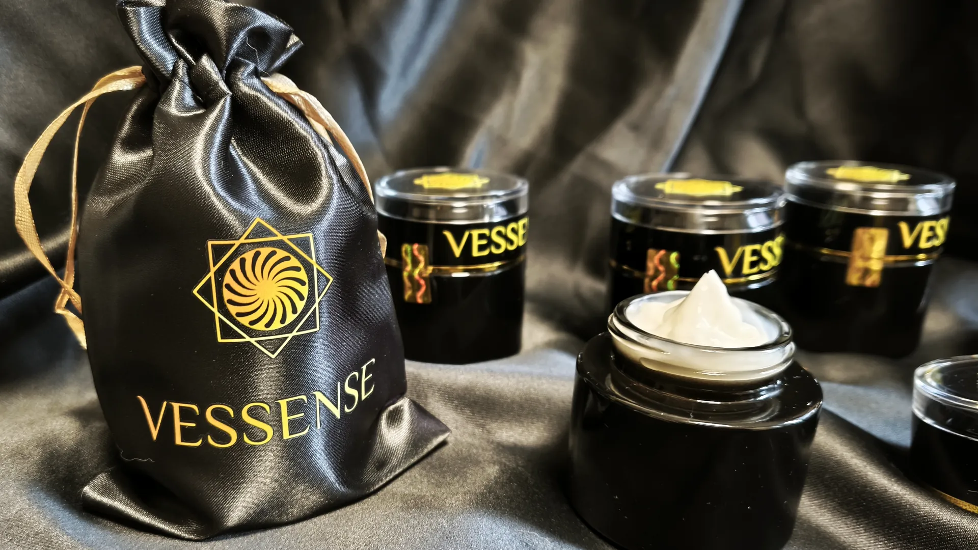 VESSENSE collection on black satin — satin pouch, sealed jars and an open jar of cream